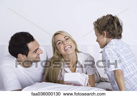 Stock Image - Family lying in bed together, woman holding gift and smiling at camera. Fotosearch