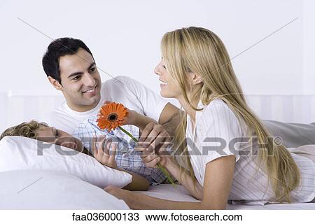 Stock Image - Family relaxing together on bed, woman holding flower. Fotosearch