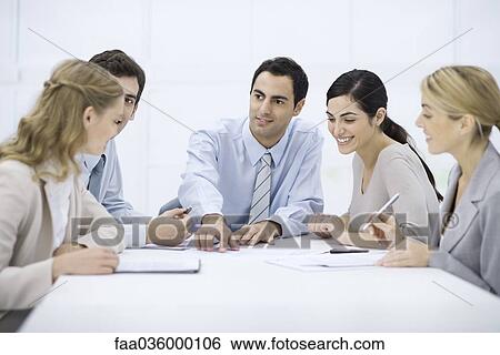 Stock Photograph - Group of professionals sitting at table, listening to male colleague pointing at document. Fotosearch