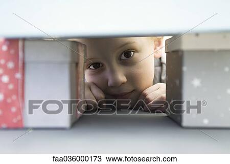 Stock Image - Little boy hiding behind pile of presents, peeking at camera. Fotosearch
