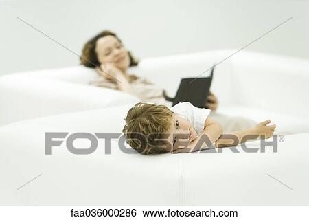 Little boy reclining in armchair, looking at camera, mother lying on sofa in background View Large Photo Image Stock Photograph - Little boy reclining in armchair, looking at camera, mother lying on sofa in background. Fotosearch