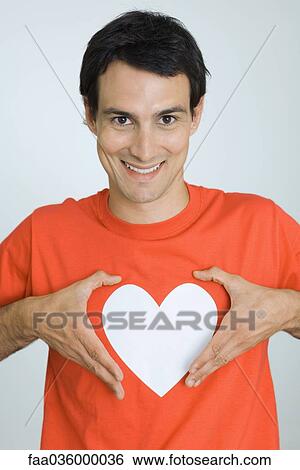 Man holding hands around heart symbol printed on his tee-shirt, smiling at camera View Large Photo Image Stock Photograph - Man holding hands around heart symbol printed on his tee-shirt, smiling at camera. Fotosearch