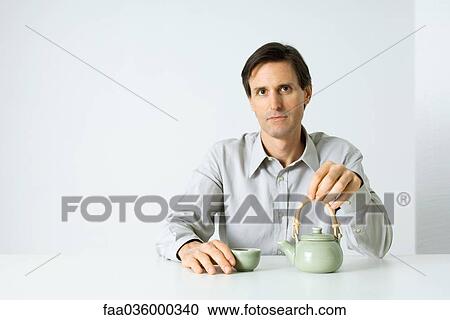 Stock Image - Man sitting with teapot and teacup, looking at camera. Fotosearch