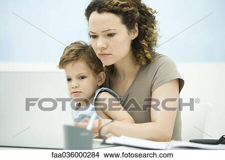 Picture - Mother holding toddler son in lap while using laptop computer. Fotosearch
