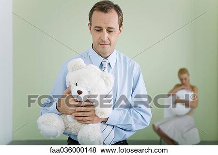 New father holding teddy bear, looking down, wife holding baby in background View Large Photo Image Stock Photo - New father holding teddy bear, looking down, wife holding baby in background. Fotosearch