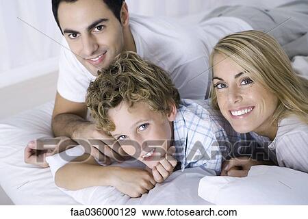 Stock Photo - Parents and son lying on bed, smiling at camera, portrait. Fotosearch