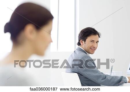 Professional man sitting at desk, smiling over shoulder at camera, female colleague in foreground View Large Photo Image Stock Photo - Professional man sitting at desk, smiling over shoulder at camera, female colleague in foreground. Fotosearch