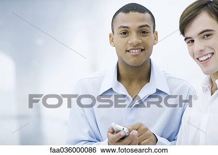 Two young businessmen smiling at camera, one holding cell phone View Large Photo Image Stock Photograph - Two young businessmen smiling at camera, one holding cell phone. Fotosearch