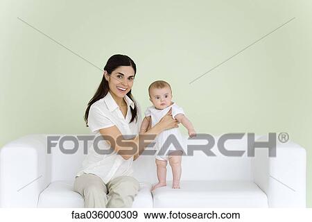 Stock Photo - Woman helping baby stand on couch, smiling at camera. Fotosearch