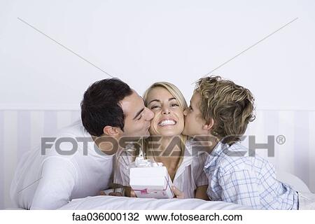 Stock Image - Woman holding a gift, husband and son kissing her on both cheeks. Fotosearch