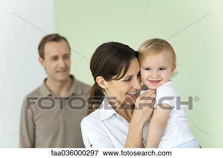 Stock Photo - Woman holding up baby, husband standing in background. Fotosearch