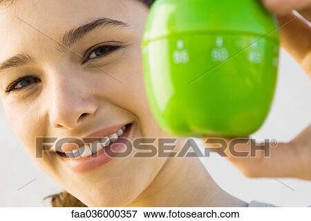 Stock Photo - Woman holding up kitchen timer, smiling at camera, focus on background. Fotosearch