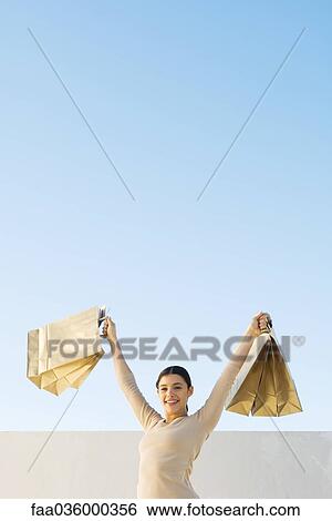 Woman holding up shopping bags, smiling at camera, low angle view View Large Photo Image Stock Photograph - Woman holding up shopping bags, smiling at camera, low angle view. Fotosearch