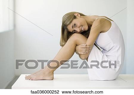 Stock Image - Woman in slip sitting on table, hugging knees, smiling at camera. Fotosearch