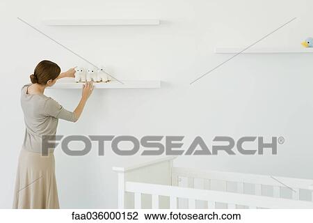 Stock Image - Woman placing stuffed toy on shelf in nursery, rear view. Fotosearch