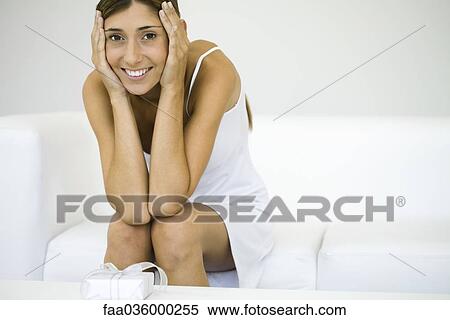 Woman sitting on sofa, holding head with both hands, gift on coffee table, smiling at camera View Large Photo Image Stock Photography - Woman sitting on sofa, holding head with both hands, gift on coffee table, smiling at camera. Fotosearch