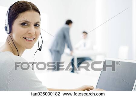 Stock Image - Woman wearing headset, working at desk, smiling over shoulder at camera. Fotosearch