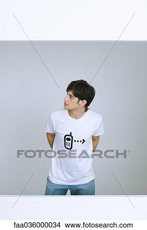 Young man wearing tee-shirt with cell phone graphic, looking away, hands behind back View Large Photo Image Picture - Young man wearing tee-shirt with cell phone graphic, looking away, hands behind back. Fotosearch