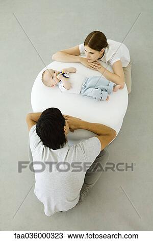 Young parents leaning against ottoman, looking at baby lying between them View Large Photo Image Stock Image - Young parents leaning against ottoman, looking at baby lying between them. Fotosearch