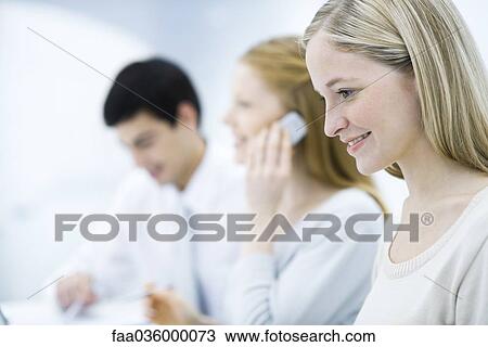 Stock Image - Young professional woman looking down, colleagues working in background. Fotosearch