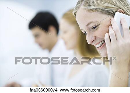 Young professional woman talking on cell phone, colleagues working in background View Large Photo Image Picture - Young professional woman talking on cell phone, colleagues working in background. Fotosearch