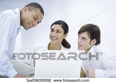 Young professionals sitting around laptop computer, leaning close together, smiling View Large Photo Image Stock Image - Young professionals sitting around laptop computer, leaning close together, smiling. Fotosearch