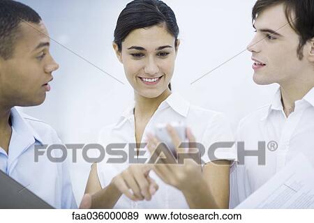 Young woman standing between two male colleagues, looking at her cell phone, smiling View Large Photo Image Stock Photo - Young woman standing between two male colleagues, looking at her cell phone, smiling. Fotosearch