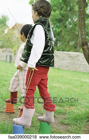 Stock Photo - Little girls using plastic buckets as stilts, side view. Fotosearch