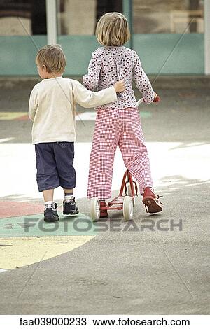 Stock Image - Siblings walking together, girl pushing scooter, boy's arm around girl's waist, rear view. Fotosearch