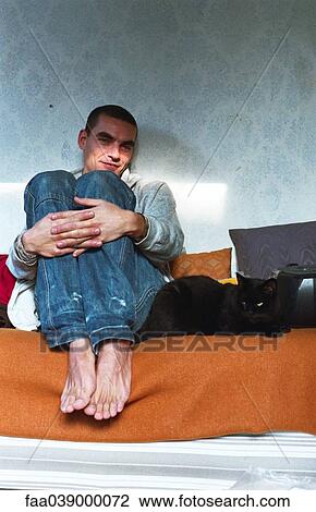 Stock Image - Young man sitting in bed hugging knees, cat lying alongside. Fotosearch
