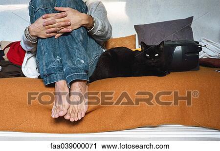 Stock Image - Young man sitting on bed hugging knees, cat lying alongside . Fotosearch
