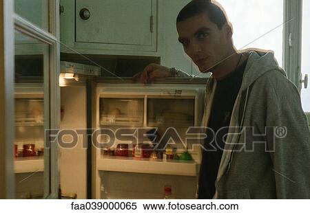 Stock Photography - Young man standing in front of open refrigerator, looking at camera. Fotosearch