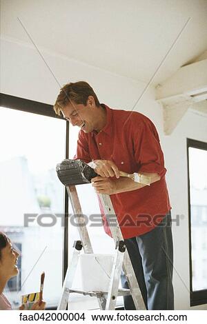 Picture - "Couple holding paintbrushes, laughing, man standing on ladder". Fotosearch