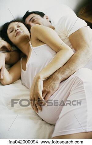 Picture - "Couple lying together on bed, both touching woman's pregnant belly". Fotosearch
