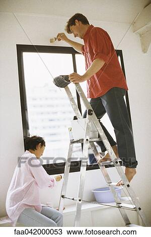 Stock Image - "Couple painting wall together, man standing on ladder". Fotosearch