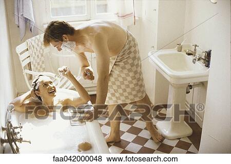 Stock Photo - "Couple together in bathroom, woman in bathtub, man leaning over her with shaving cream on face". Fotosearch