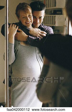 Stock Image - Expecting couple embracing in front of mirror. Fotosearch