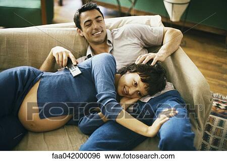 Stock Photograph - "Expecting couple relaxing on sofa, watching TV, woman resting head in man's lap". Fotosearch