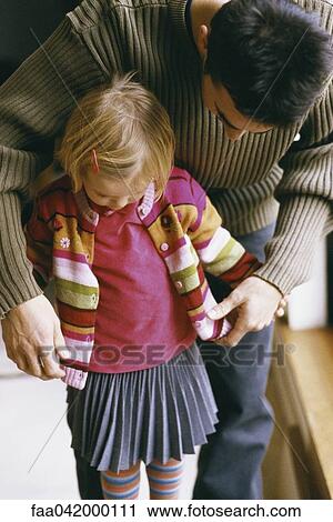 Stock Image - Father bending over to help young daughter button cardigan. Fotosearch