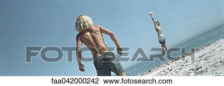 Stock Image - "Friends throwing frisbee at beach, one jumping to catch frisbee". Fotosearch