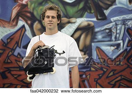 Stock Image - Male with pair of inline skates hanging over shoulder standing in front of wall mural. Fotosearch