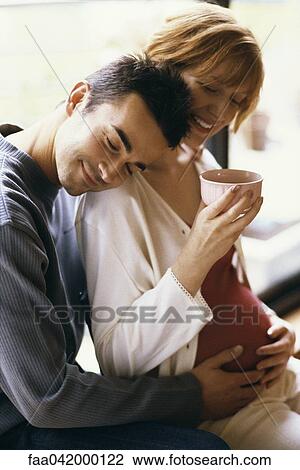 Stock Image - "Man embracing pregnant wife, hands on her stomach, woman drinking tea". Fotosearch