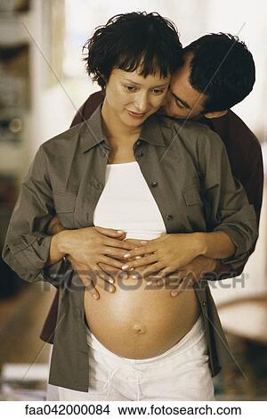 Picture - "Man embracing pregnant woman from behind, both touching her stomach". Fotosearch