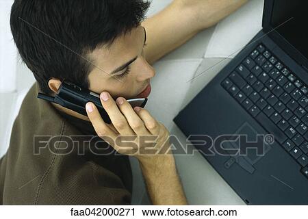Stock Image - "Man phoning, leaning on elbow, using laptop computer". Fotosearch