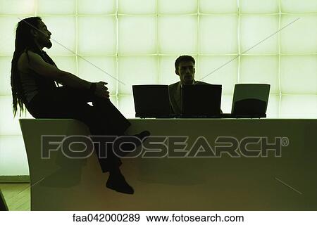 Stock Photo - "Man with dreadlocks sitting on desk with legs crossed, speaking to businessman using laptop computer". Fotosearch