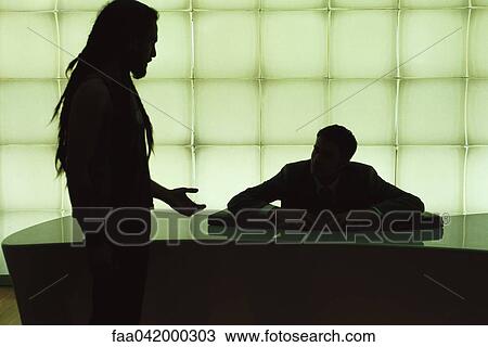 Stock Image - Man with dreadlocks speaking to businessman seated behind desk. Fotosearch