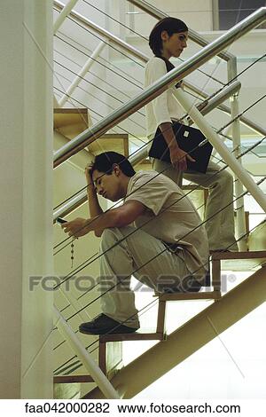 Stock Image - "Man with hand on head sitting on stairs looking at cell phone, woman walking past". Fotosearch