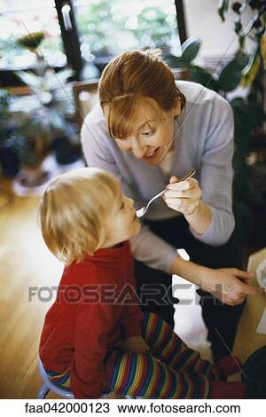 Stock Image - "Mother feeding daughter with spoon, smiling". Fotosearch