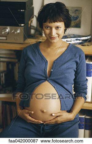 Stock Photo - "Pregnant woman holding stomach, smiling at camera". Fotosearch
