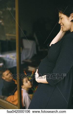Stock Photograph - "Pregnant woman leaning in doorway, smiling, family in background". Fotosearch